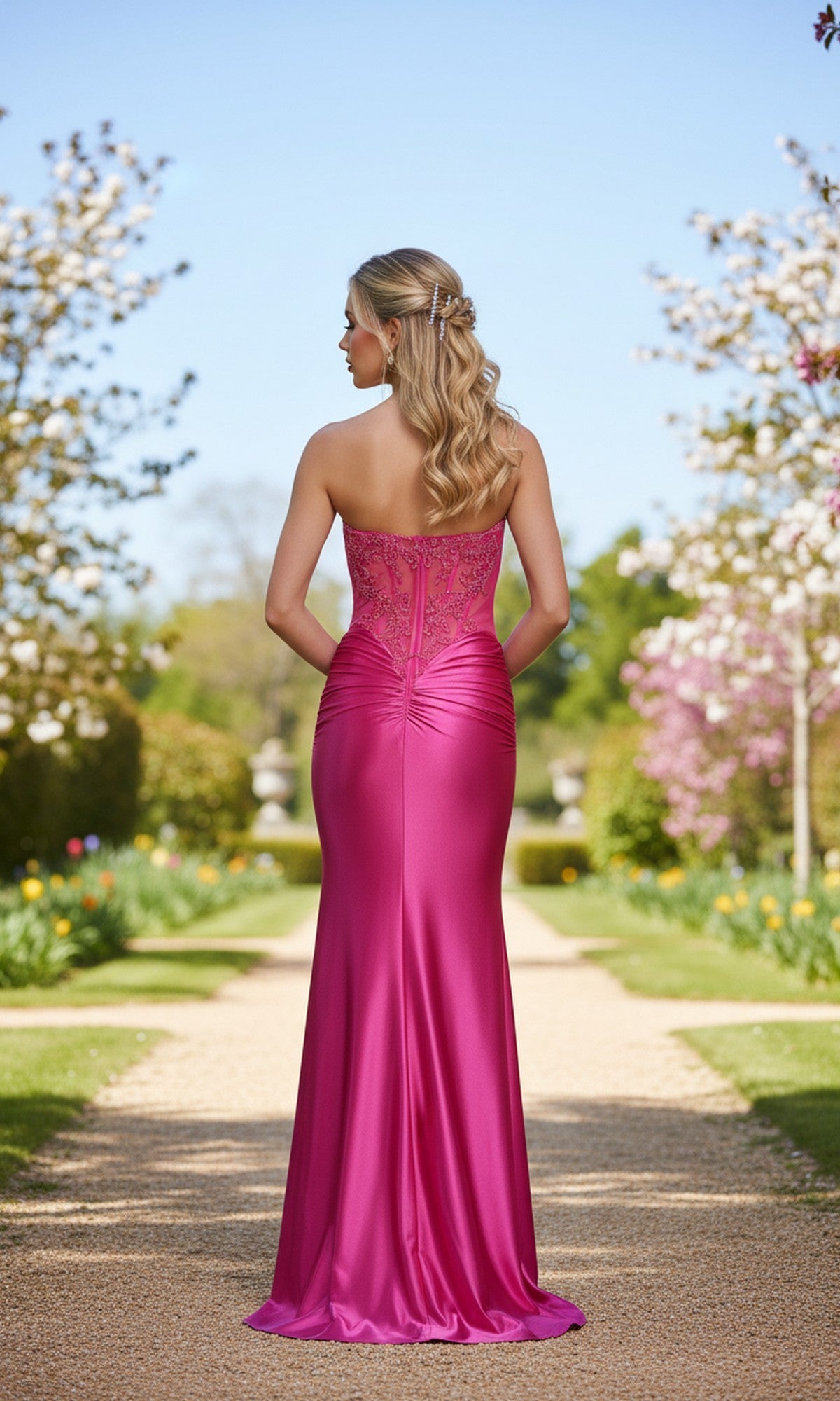 A woman with long blonde hair wears the La Femme 33639 long prom dress with a corset bodice, standing on a garden path amid blooming trees, facing away from the camera.